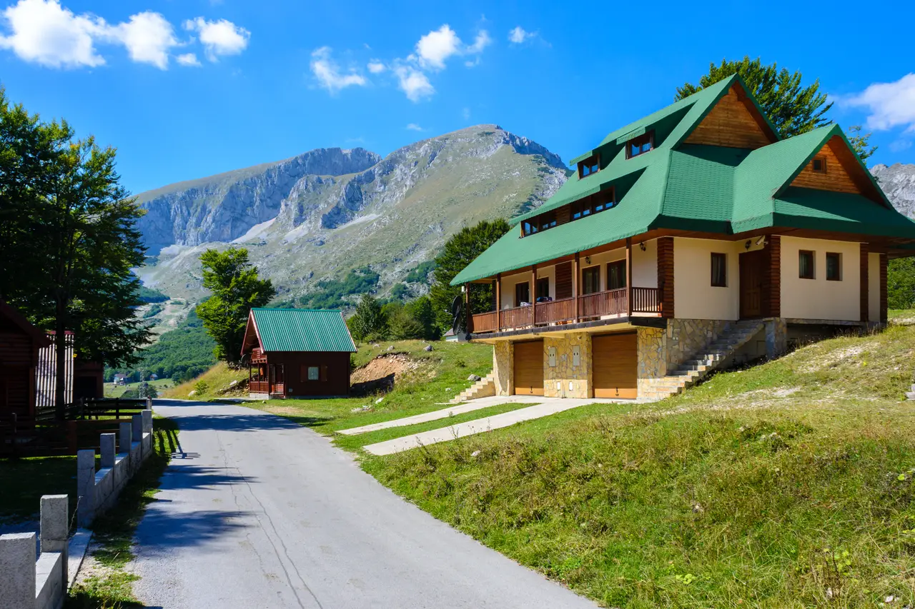 A large timber guesthouse with a green roof and balcony beside a quiet lane, a jagged Durmitor ridge behind