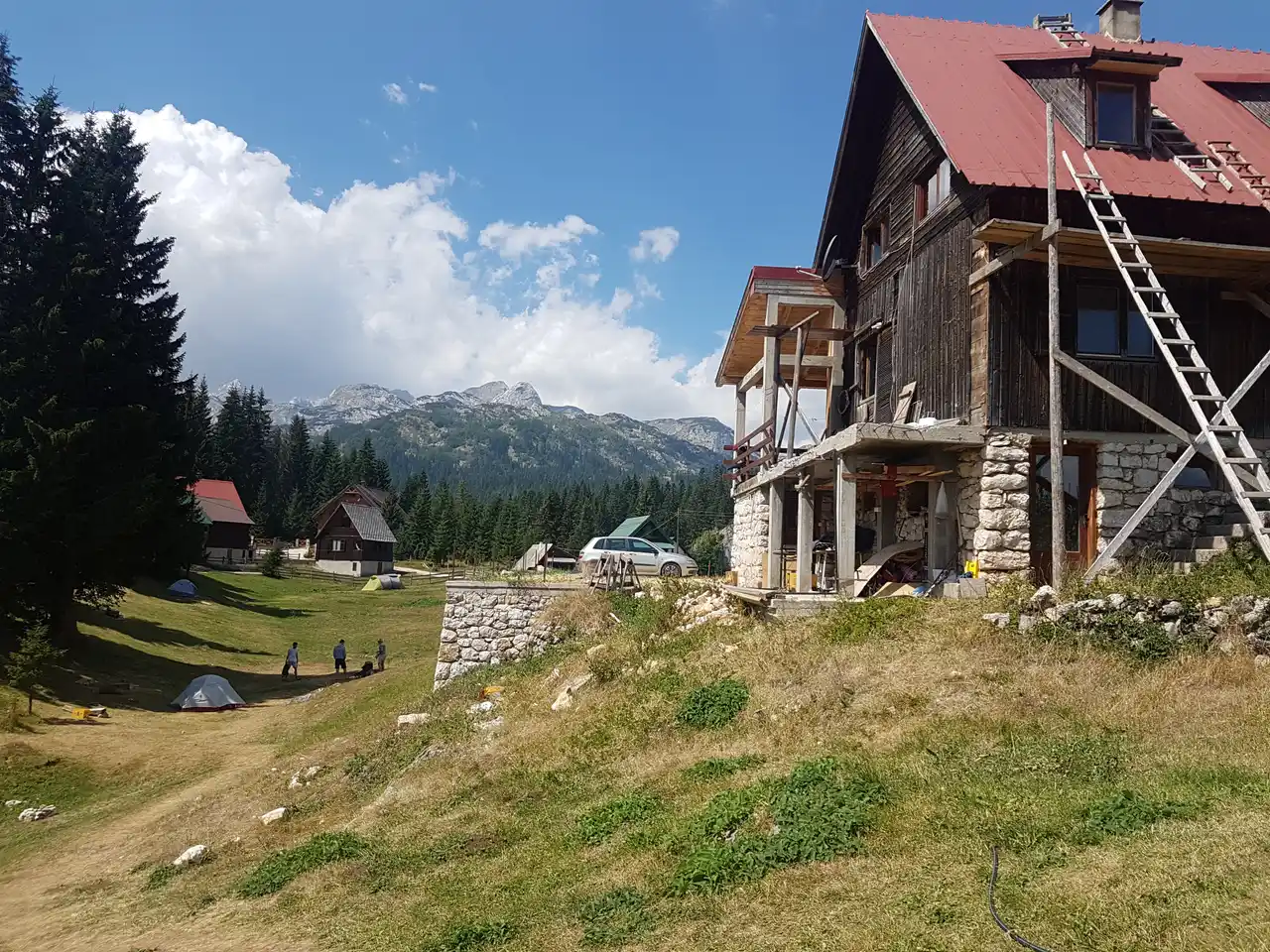 A timber mountain lodge with a red roof beside a grassy camping area with tents and pine trees, Durmitor peaks behind