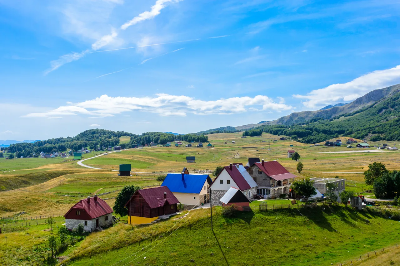 A cluster of houses with red and blue roofs scattered across the green Žabljak plateau, mountains behind
