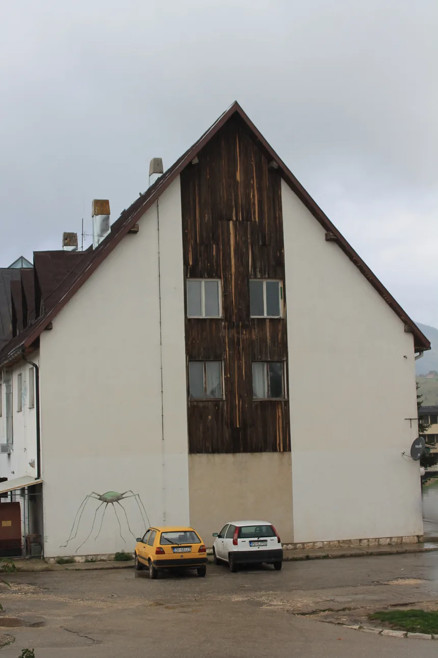 A tall town building in Žabljak with a dark timber gable and two small cars parked below under a grey sky